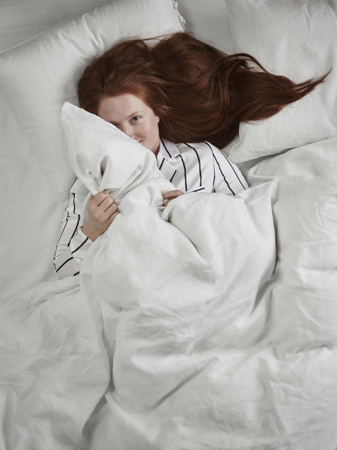A woman with red hair laying in a bed with a white comforter and white pillows.