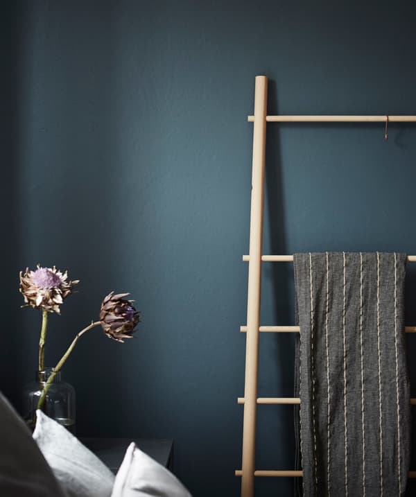 A wooden blanket ladder leans against a dark blue/green wall in a bedroom.