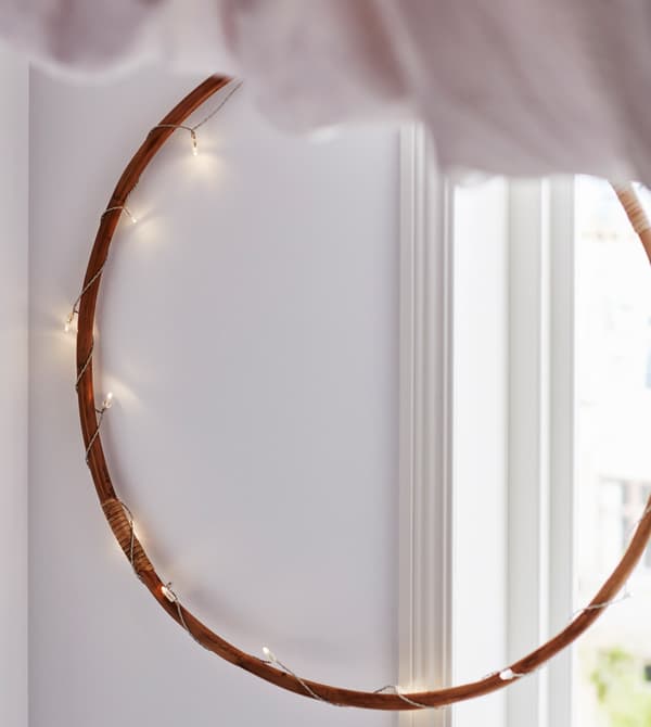 A wooden hoop is wound with a string of lights and hangs on the wall, partly infront of a window.
