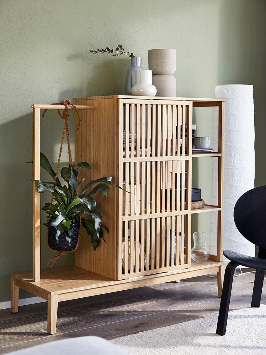 A wooden shelving unit with slatted doors, featuring potted plants and decorative vases, against a light green wall.