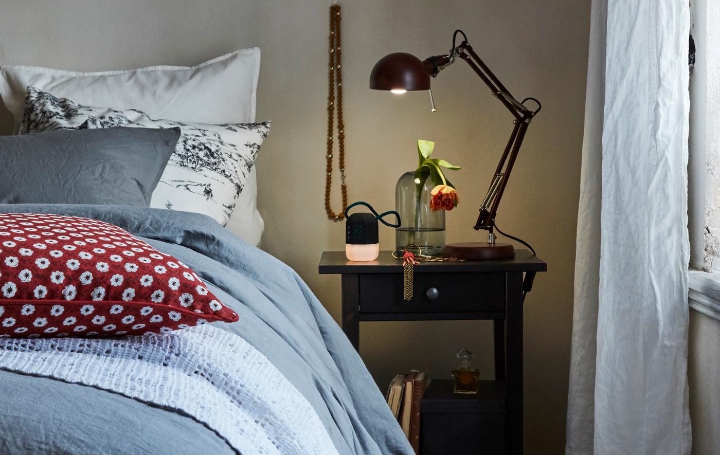 A work lamp on a table next to a bed shines light on a glass vase and alarm clock in a cosy bedroom.