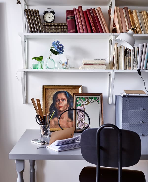A workspace in a white dorm room, including a black office chair, grey desk, artwork, and wall shelves with books.