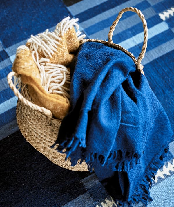 A woven basket containing a yellow fringed throw and a blue linen throw on a blue stripy patterned rug.