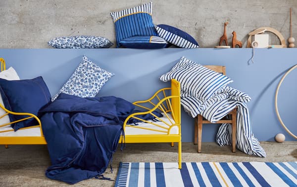 A yellow bed frame with blue and white bedding in floral and striped prints, and cushions and wooden toys on a blue wall.