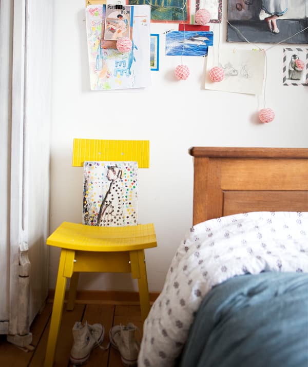 A yellow chair against a white wall decorated with pictures and notes, next to a wooden bed.