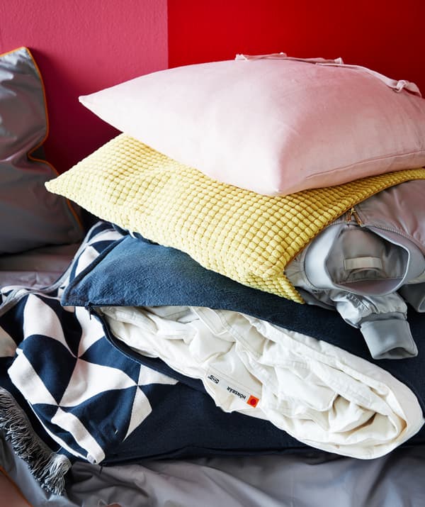 A yellow GULLKLOCKA cushion cover stuffed with a jacket, and a blue cushion cover filled with a blanket för seasonal storage.