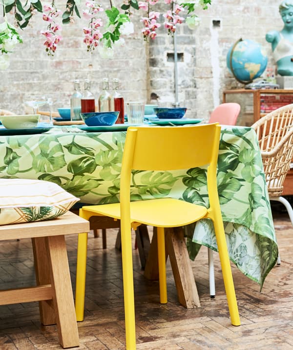 A yellow plastic dining chair pulled up to a table covered with a green leaf-print tablecloth and blue crockery.