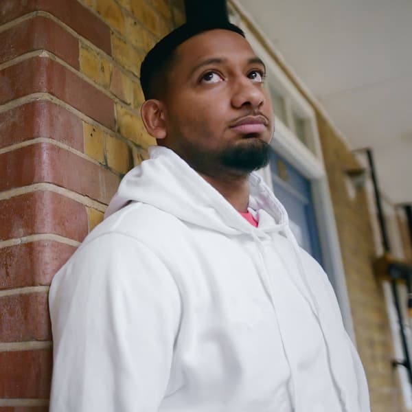 A young bearded man in a white hoodie leaning against a brick wall while looking towards the sky.