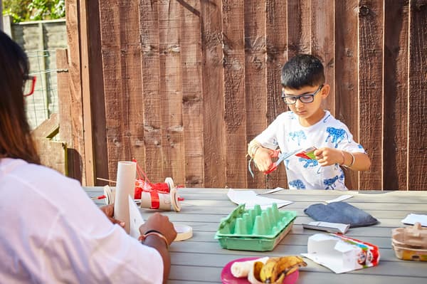 A young boy is cutting cardboard with a pair of red scissors by a wooden table.