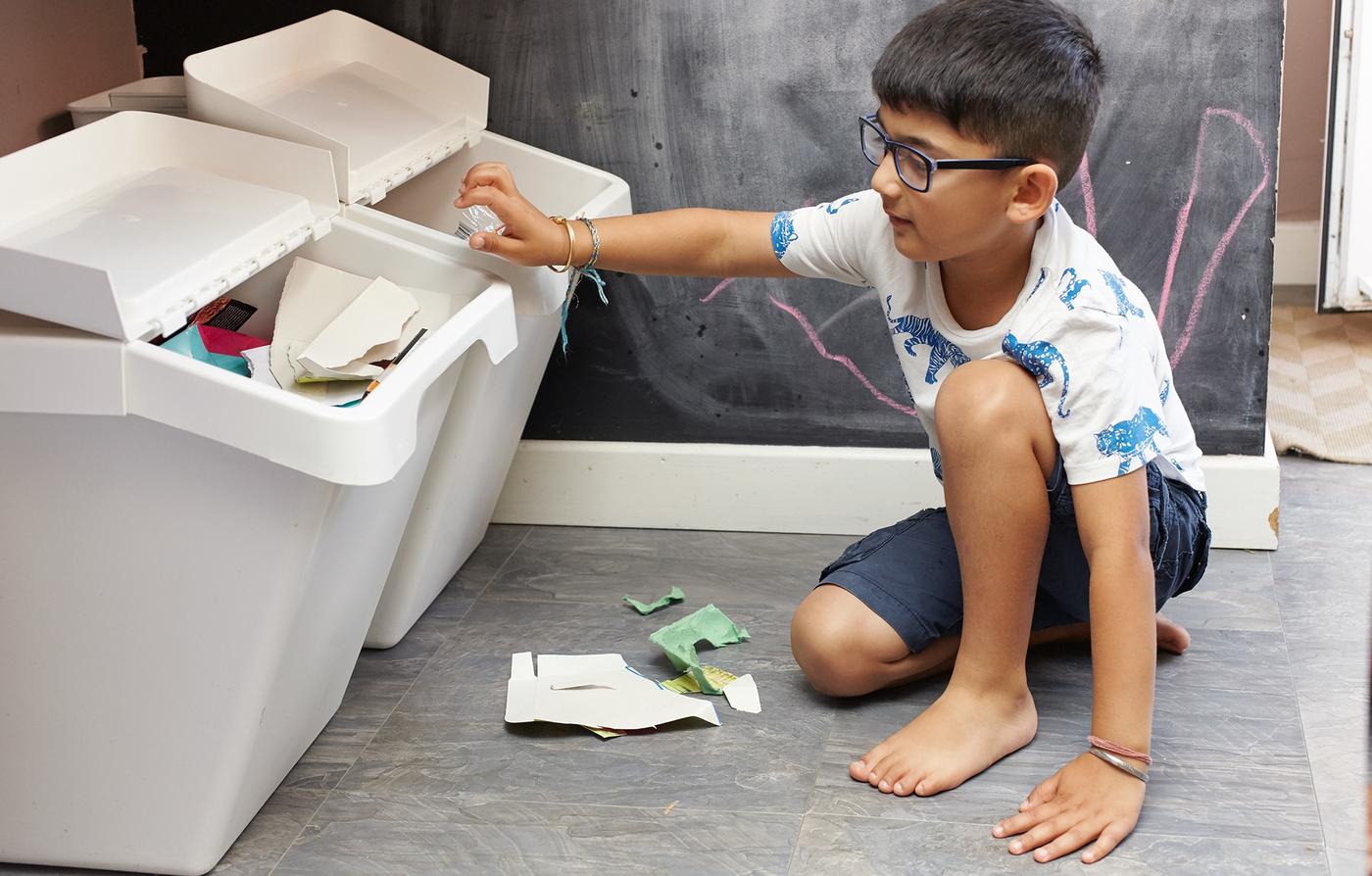 A young boy is sitting on the floor and recycling cardboard in white sorting bins.  