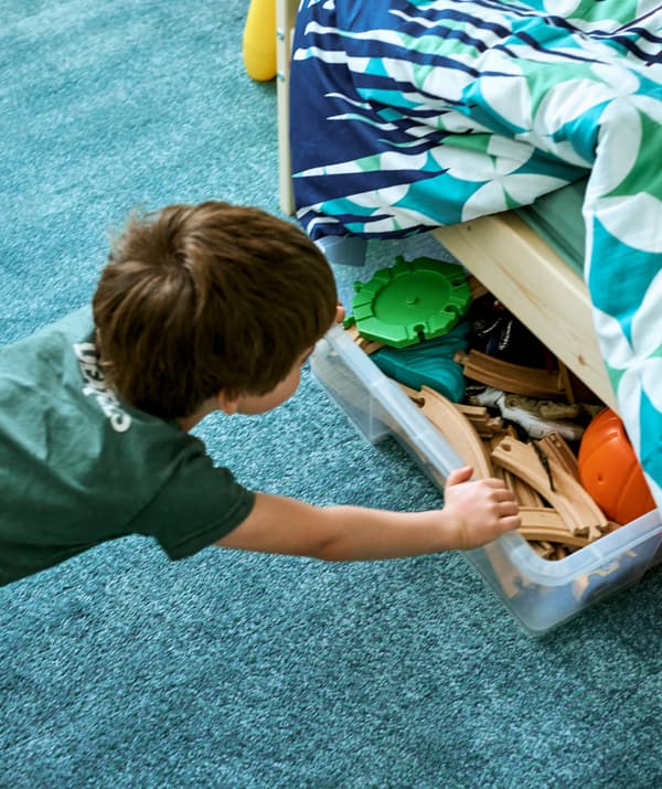 A young boy pushes a clear storage box full of toy train track pieces under a bed with colorful bedding on a turquoise rug.