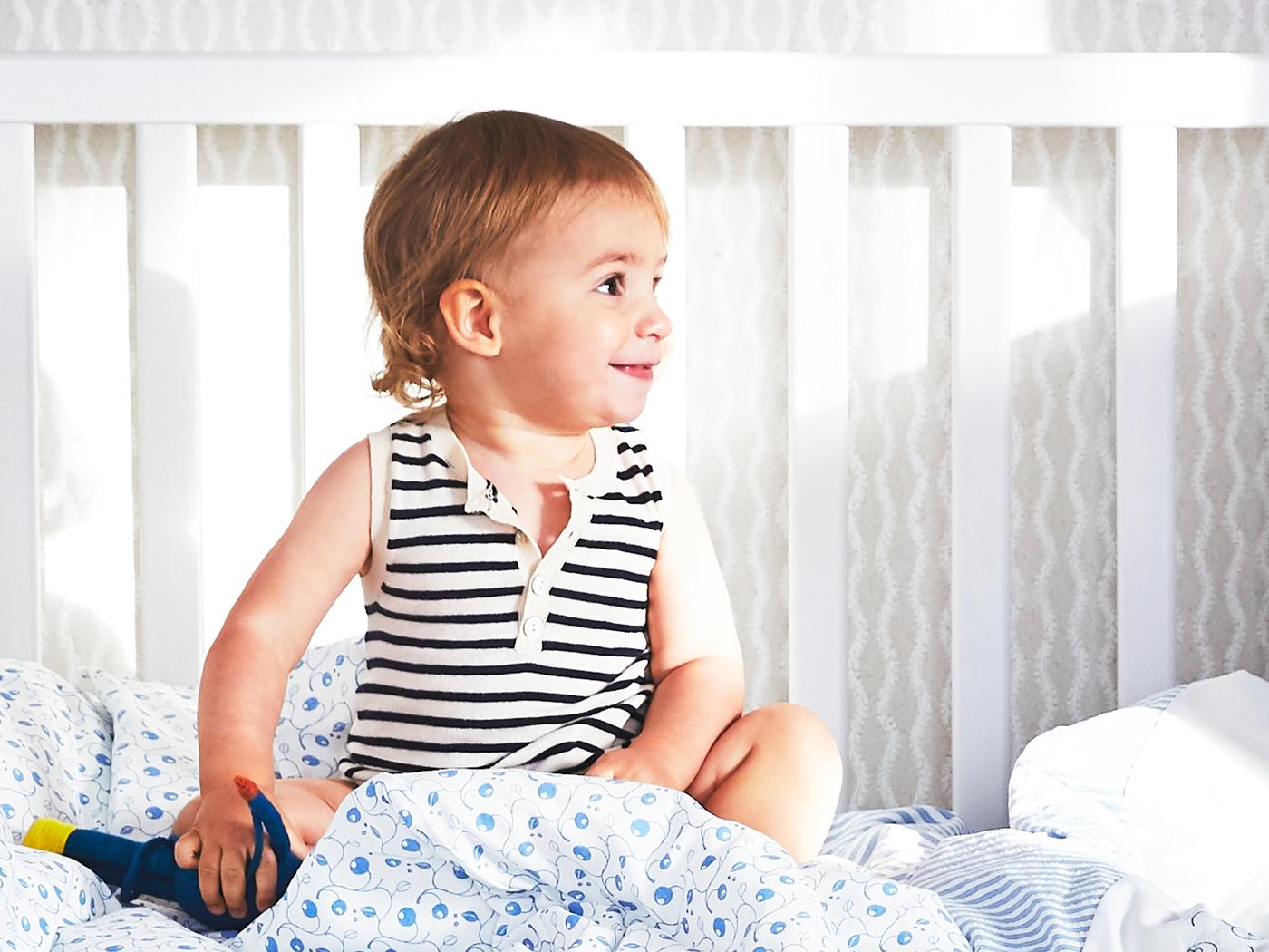 A young child sits in a white SUNDVIK cot with one side removed and GULSPARV bed linen with a blueberry pattern.