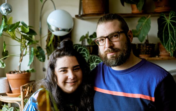 A young couple in their urban apartment with lots of plants behind them.