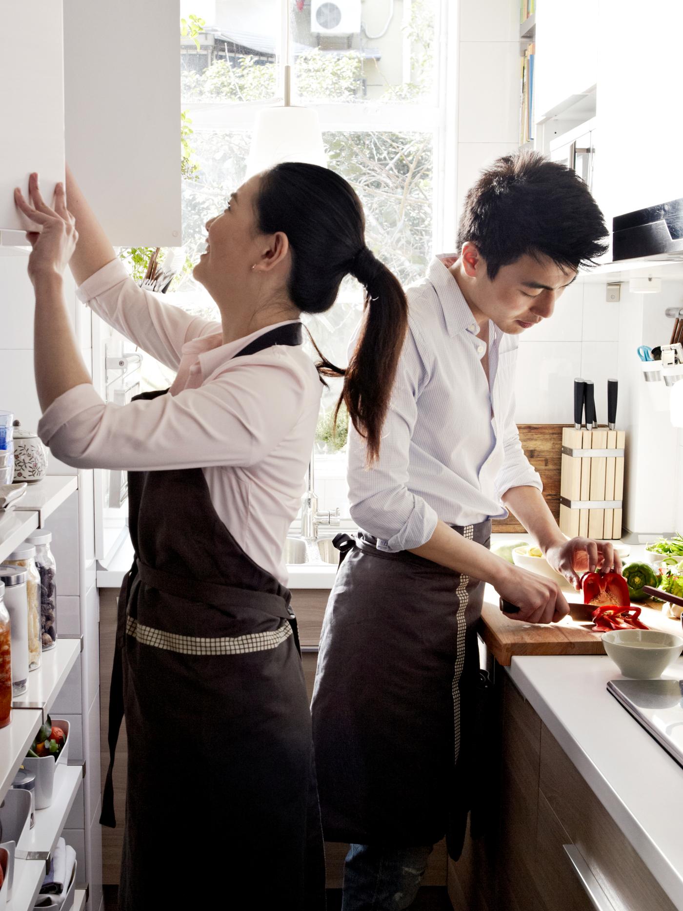 A young couple preparing food in their galley kitchen. One of them chops a pepper while the other looks in a wall cabinet.