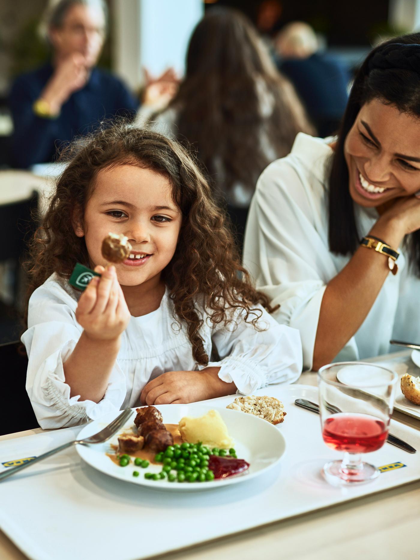 A young girl enjoys a serving of plant ball, mashed potatoes and peas in an IKEA restaurant while her mother looks on.