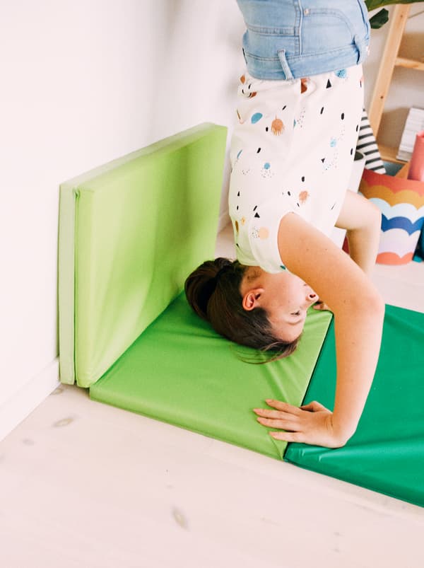 A young girl performs a handstand against a wall, her head and arms supported by a bright green PLUFSIG folding gym mat.