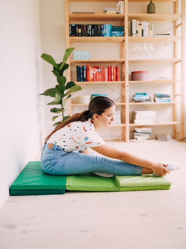 A young girl stretches while sat on a green PLUFSIG folding gym mat, with a plant and IVAR shelf in the background.