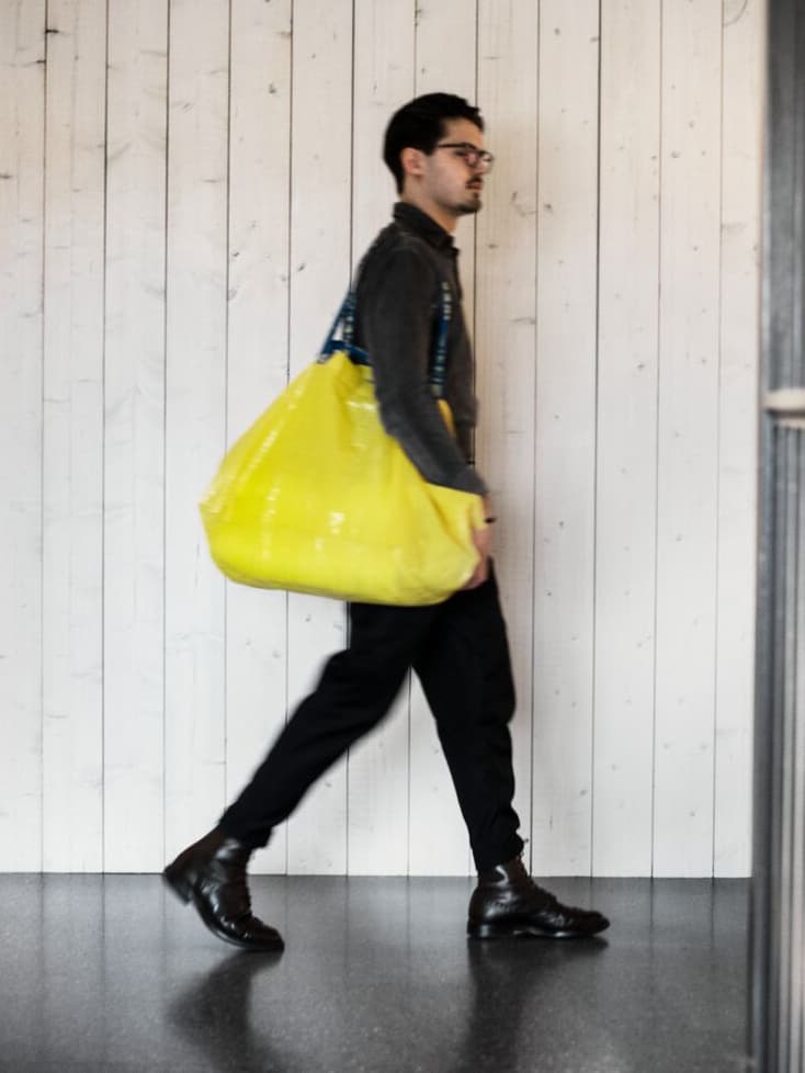 A young man dressed in black walking around in an IKEA store carrying a yellow shopping bag.