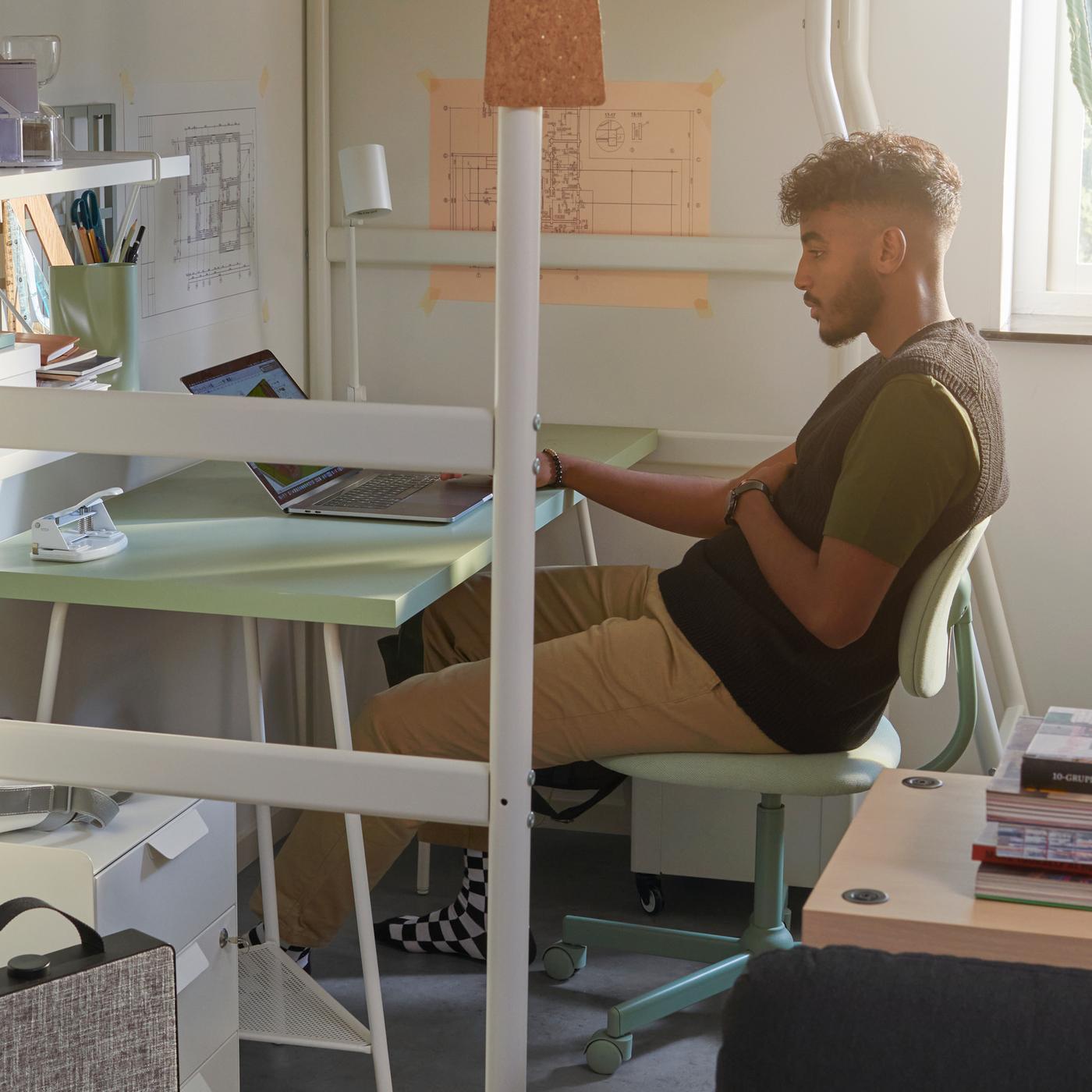 A young man sits on a BLECKBERGET swivel chair at a LAGKAPTEN desk, placed underneath a VITVAL loft bed.
