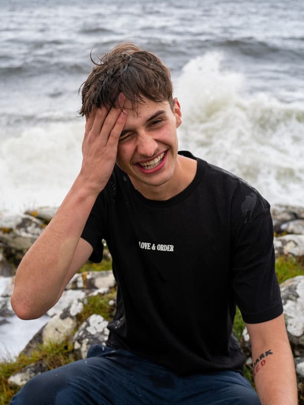 A young man wearing a black t-shirt is sitting by the ocean on a rock, laughing. Waves are crashing behind him.