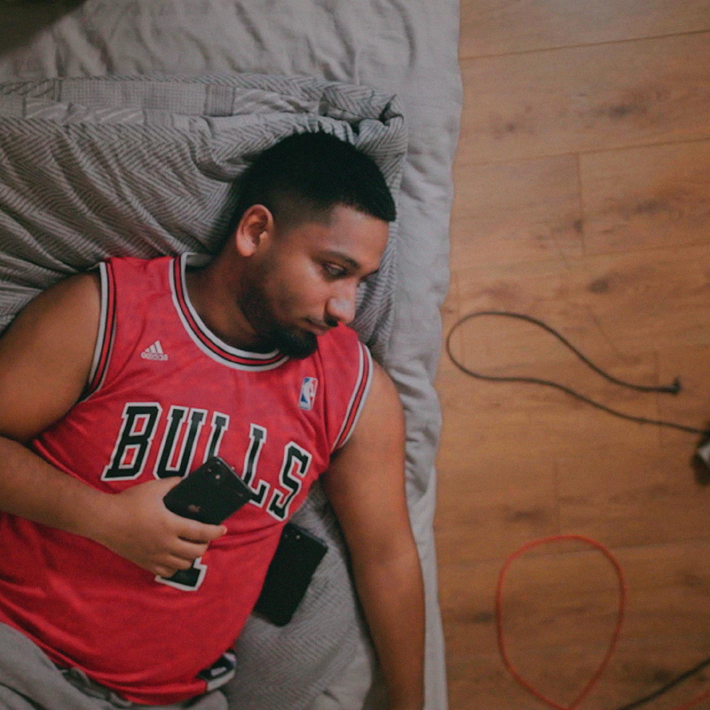 A young man with a beard and a red sports shirt is relaxing on a grey sofa while holding a phone.