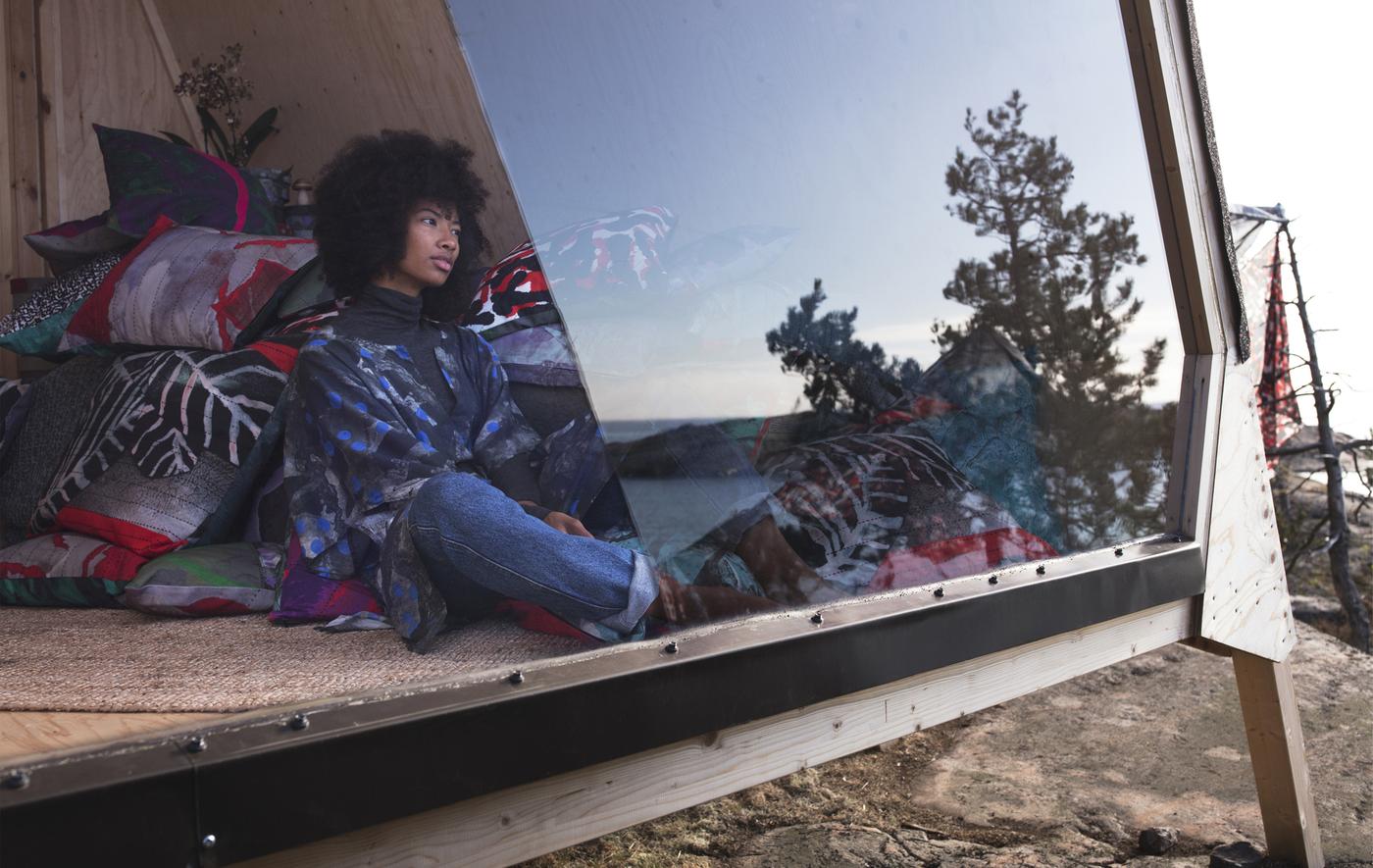 A young woman is sitting against a pile of patterned cushions, looking out of an angular window at an ocean view.