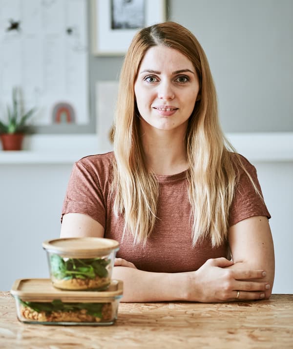 A young woman sits at a wood table with two bamboo-lidded glass food jars containing lentil stew and spinach in front of her.