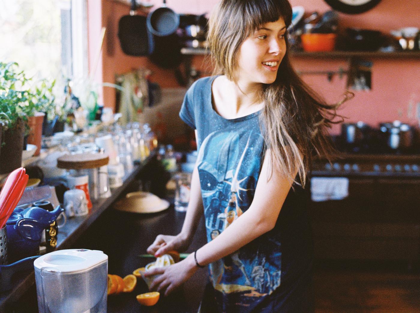A young woman turning to look at someone while she washes and cuts up pieces of fruit by the kitchen sink.