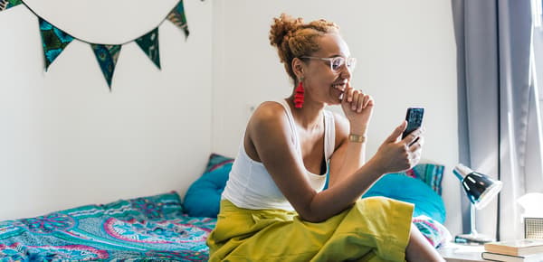 A young woman wearing bright yellow trousers and a pair of glasses is sitting on her bed. She is smiling at her phone.