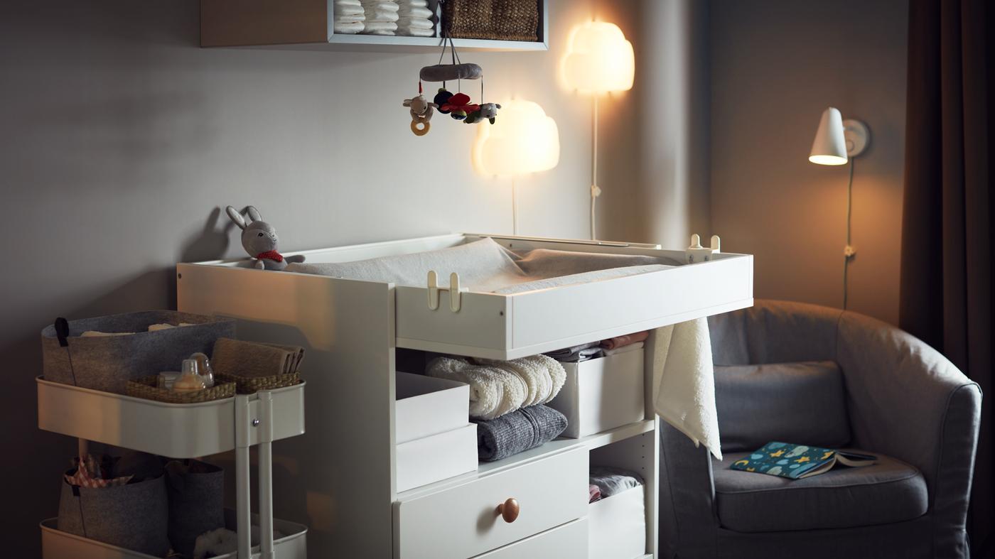 Acogedora recámara con un cambiador, un carrito de almacenamiento con accesorios para el bebé, un sillón, lámparas de pared de noche y almacenamiento de pared.