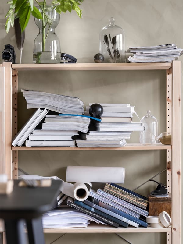 Adossé à un mur, une étagère en bois remplie de piles désordonnées de livres et de magazines avec des vases en verre ornés de plantes et de plumes.