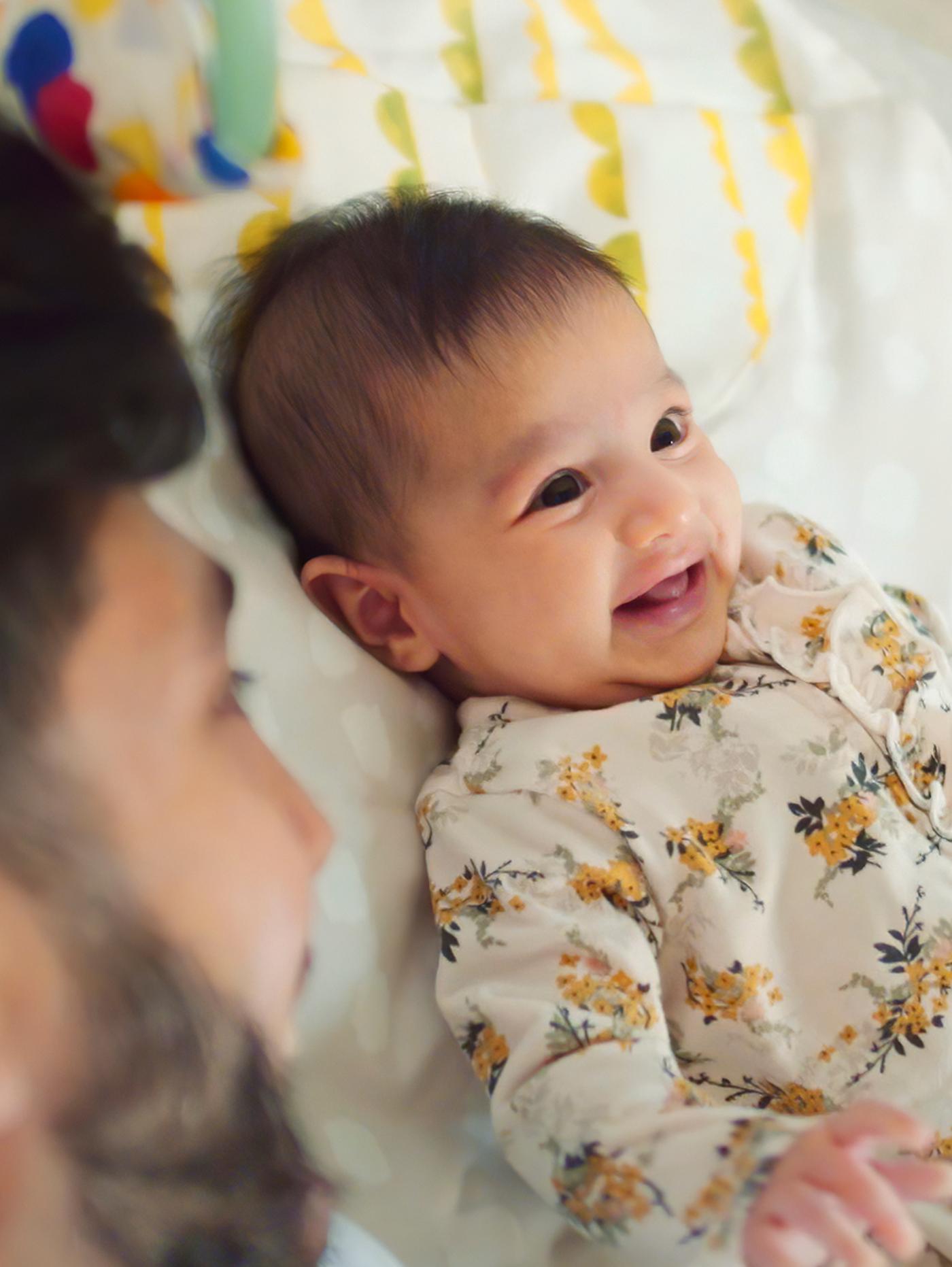 Allongé sur le dos, un bébé aux cheveux foncés vêtu d’une combinaison à fleurs sourit à son papa.