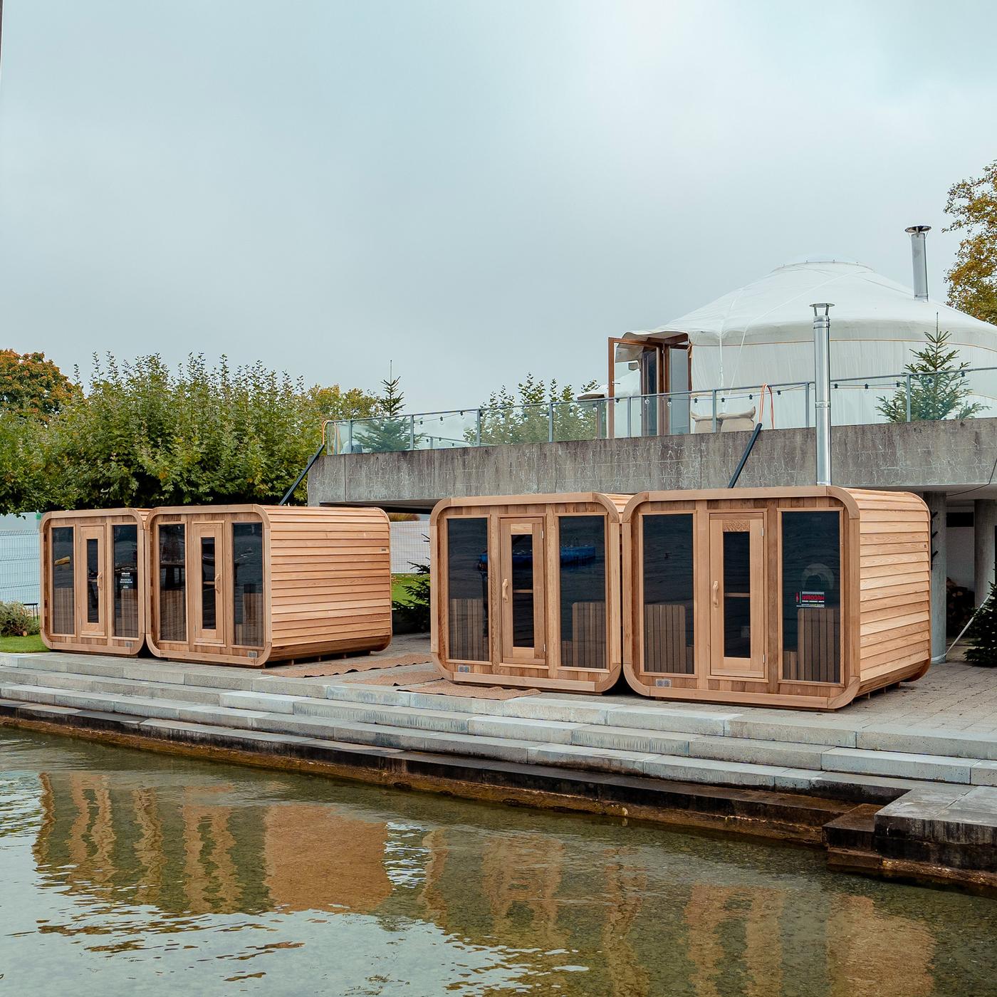 Am Rand eines Schwimmbeckens stehen vier Holz-Cubes mit je einer Sauna darin