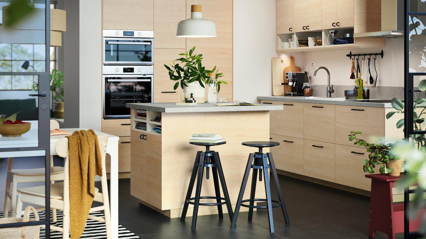 Ampia isola per cucina con frontali ASKERSUND effetto frassino chiaro e due sgabelli neri. Sopra l'isola è appesa una lampada a sospensione bianca.