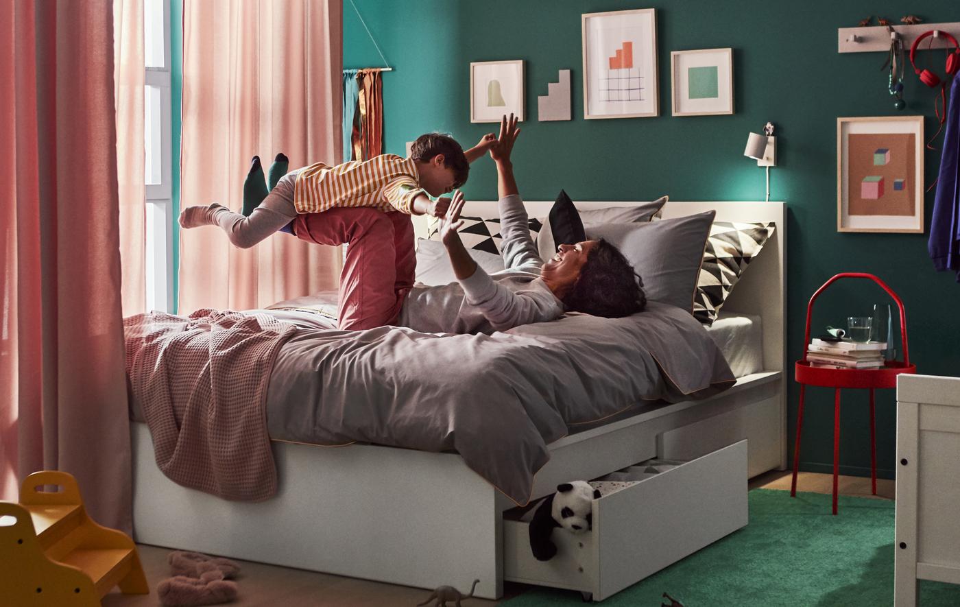 An adult and child playing on a double bed with underbed storage drawers, with curtains hanging around the bed and artwork on the walls.