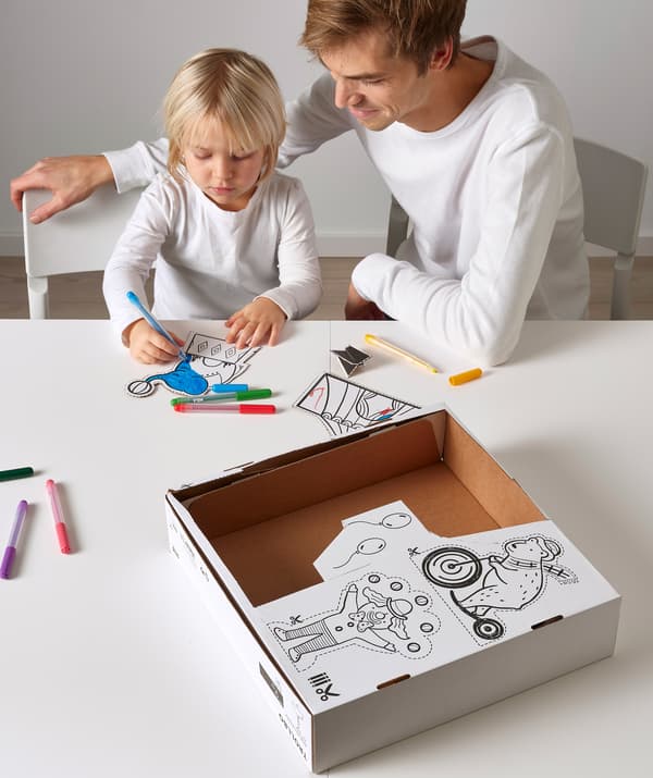 An adult sits with a child who is colouring in circus drawings, cut from the cardboard box on the table.