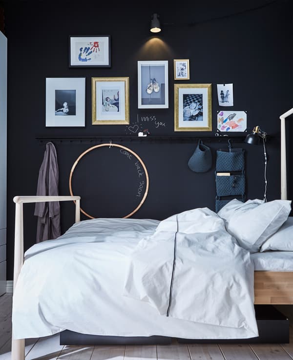 An adult’s bed in against a wall covered in children’s artwork and wall rails with hanging organisers 
