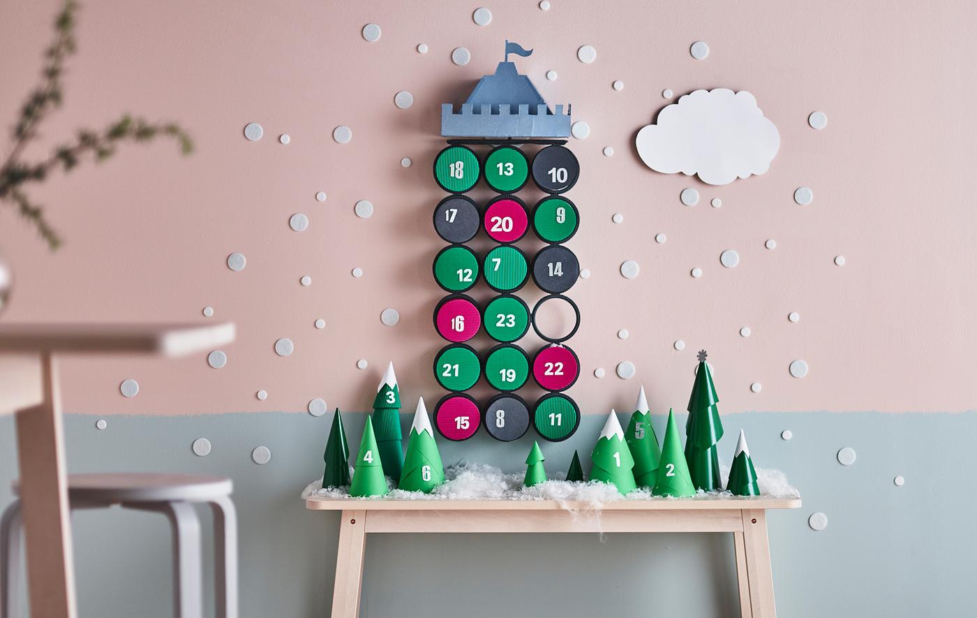 An advent calendar display with paper trees on a table and a castle hanging on a wall.