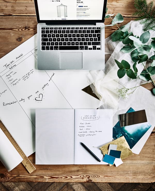 An aerial view of a desk with a computer, notebooks, a wedding planning timeline and fabric swatches