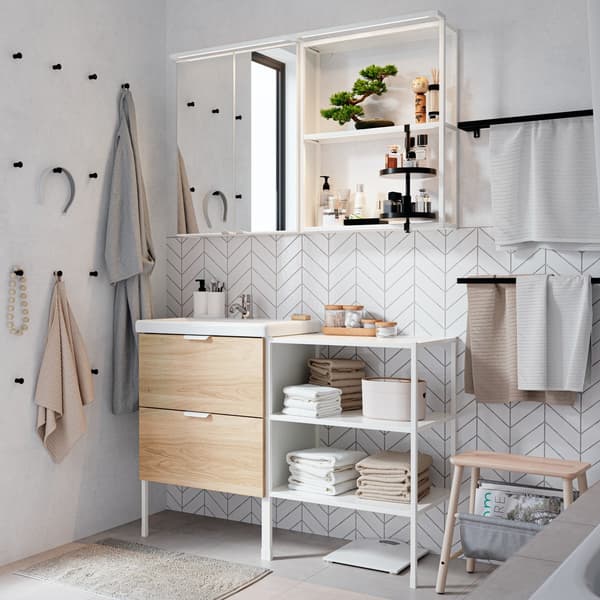 An airy, white-tiled bathroom has an oak and white ENHET/TVÄLLEN two drawer cabinet and shelves below a mirror cabinet.