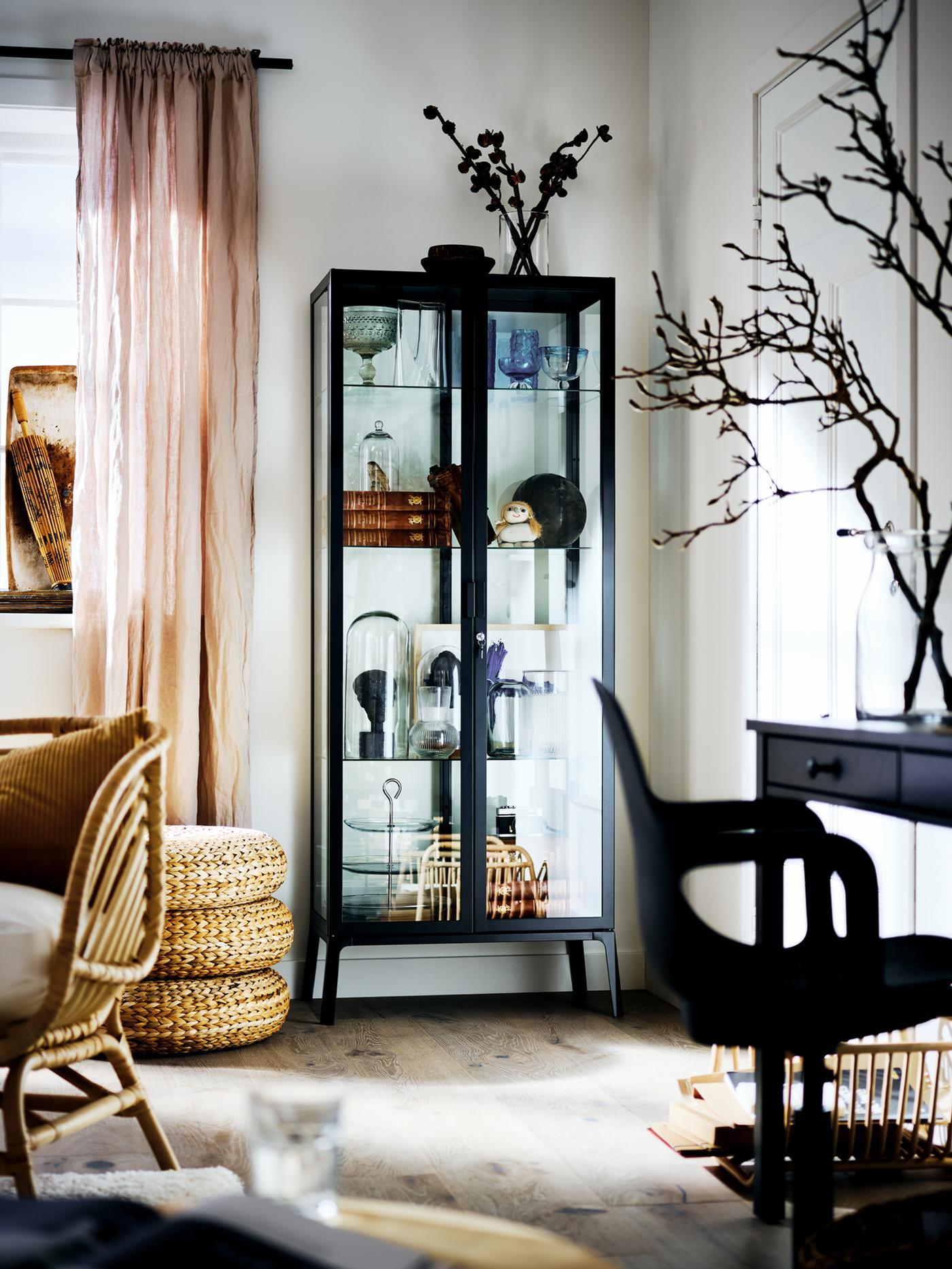 An anthracite MILSBO cabinet with glass doors filled with an assortment of objects, in the corner of a bright living room.