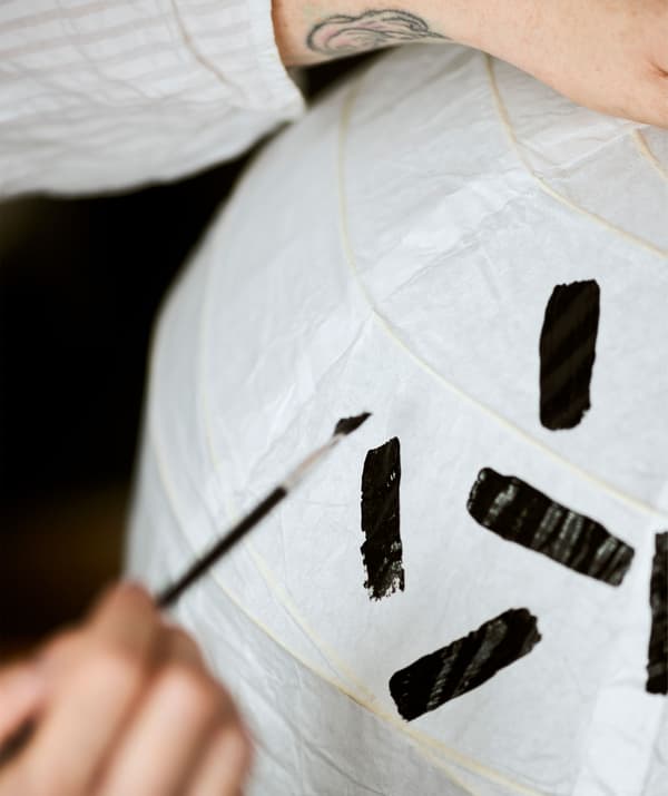 An arm with a tattooed wrist holds a white paper lamp shade steady as a pattern of black lines is painted on.