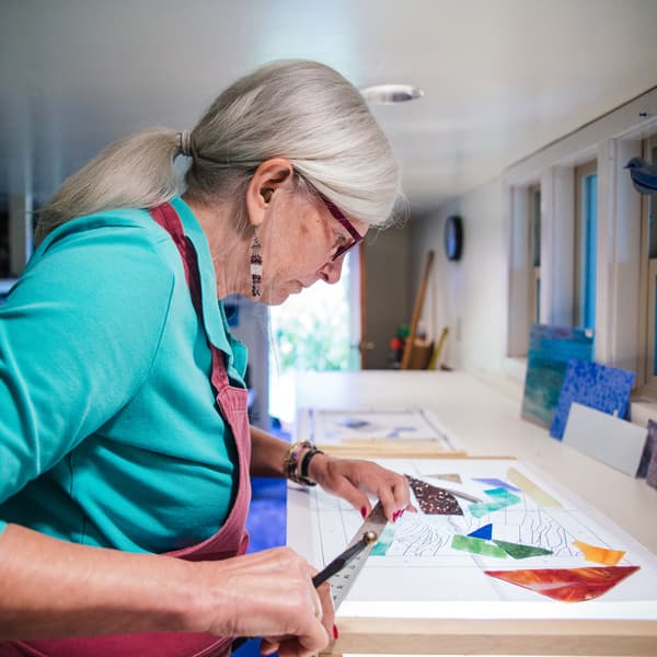 An elderly woman wearing an apron is holding a metal ruler, concentrating on the piece of art in front of her.