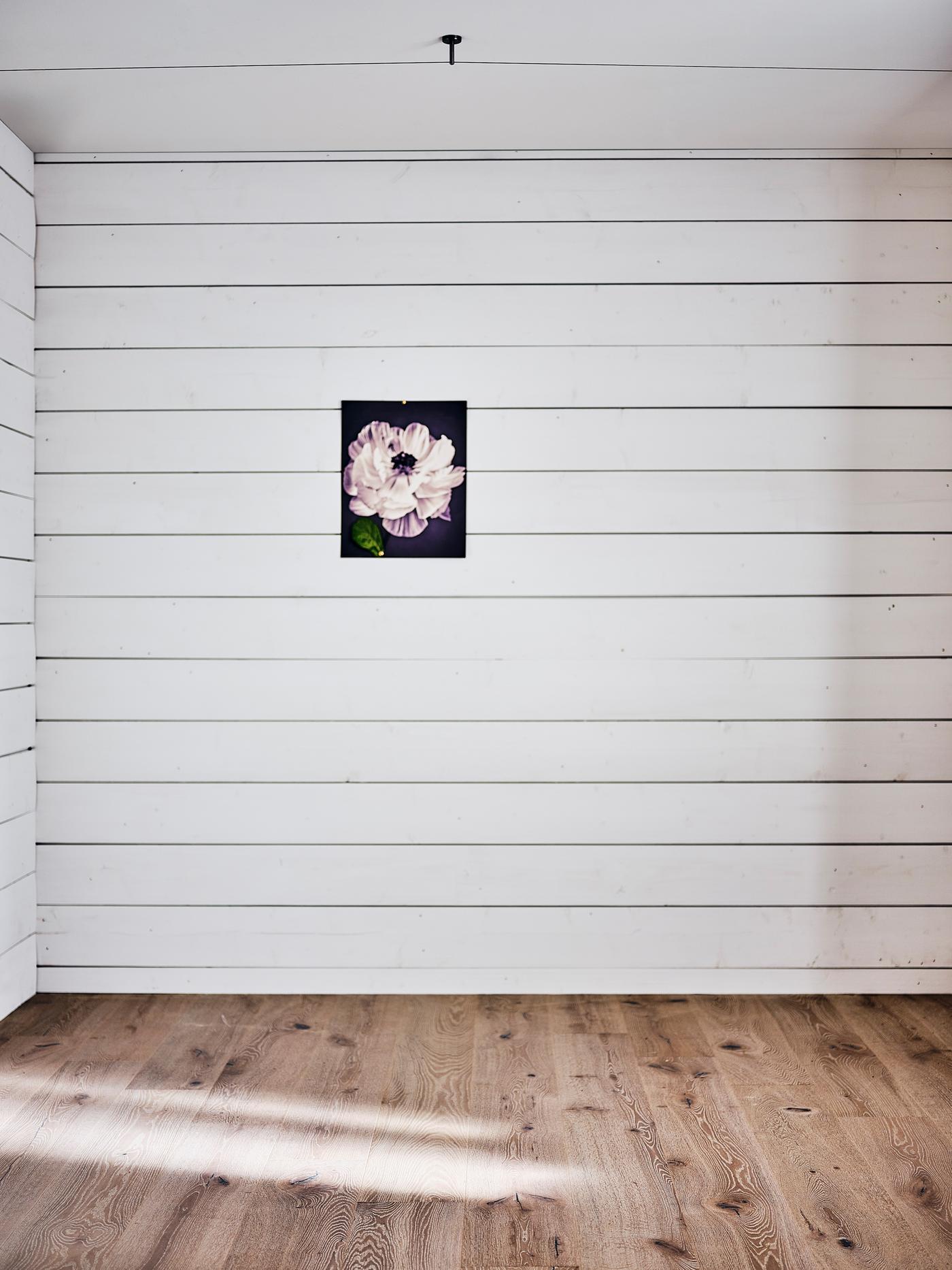 An empty bedroom with white wood-panelled walls and a light wood floor and a picture of a flower on the wall.