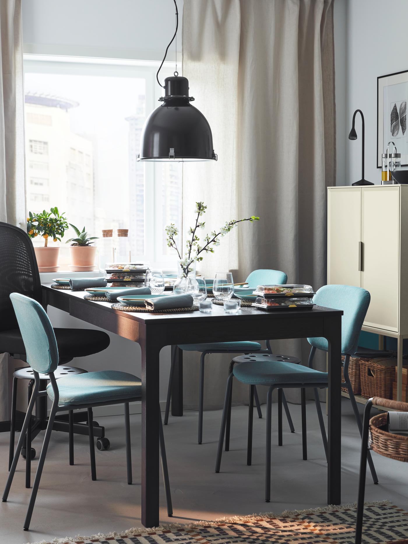 An extended brown EKEDALEN table set for dinner for six, with a mix of chairs and stools around it, beside a bright window.