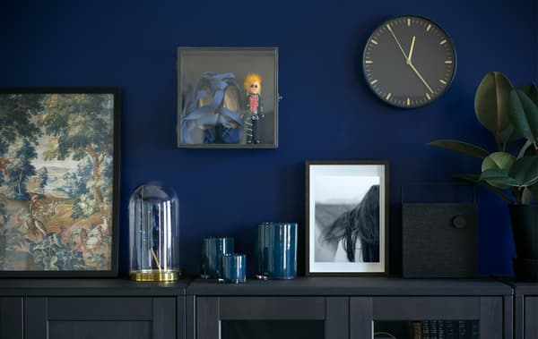An IKEA BARKHYTTAN display box on the wall, above a cabinet adorned with vases, plants and pictures.