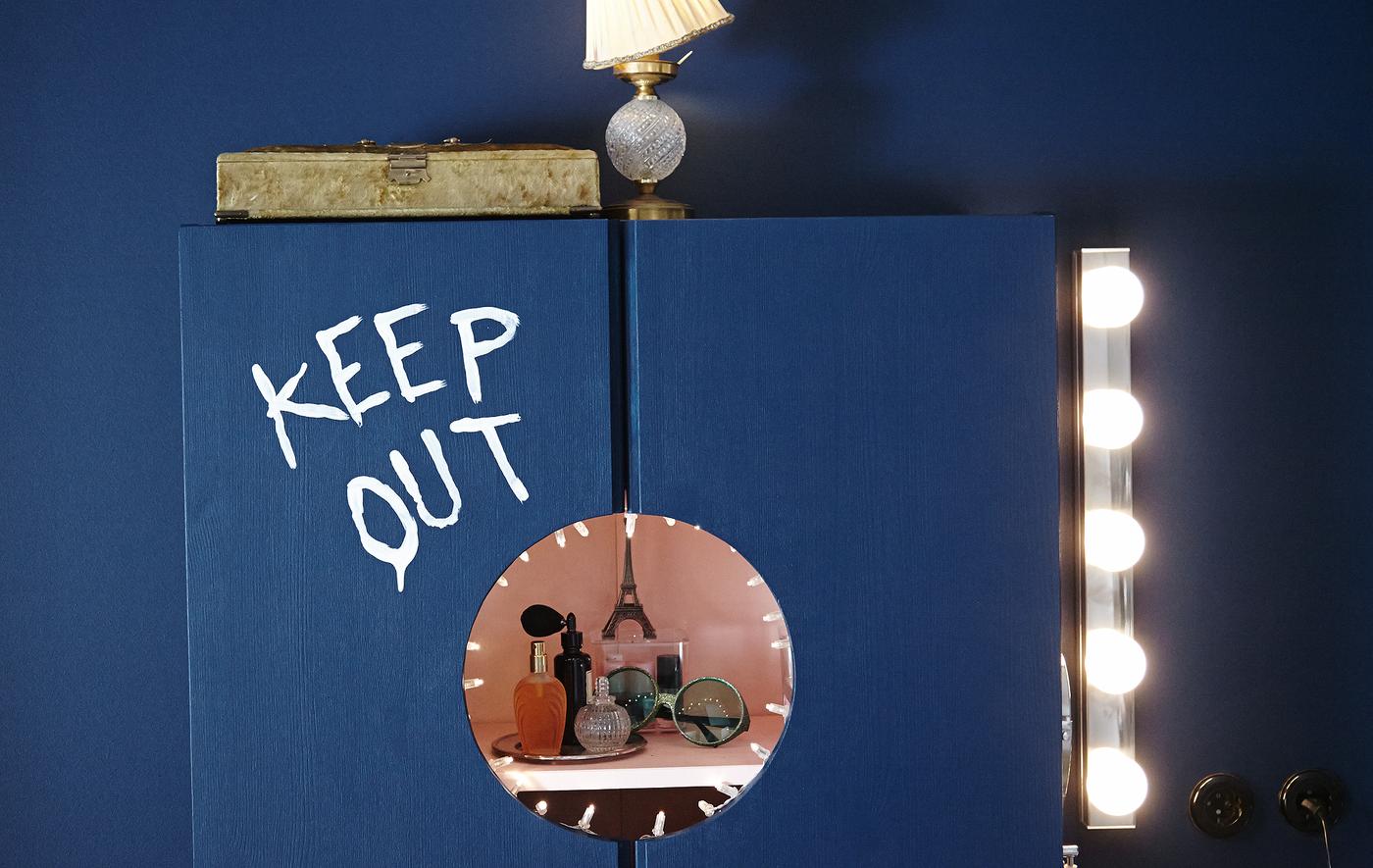 An IKEA cabinet is turned into a vanity. The words “Keep out” are painted on the front, a circle has been but from the doors for a view of the interior.