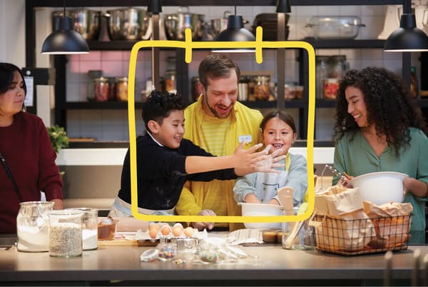 An IKEA co-worker by a kitchen counter with bowls, eggs and flour is smiling, two children and their mothers are baking.