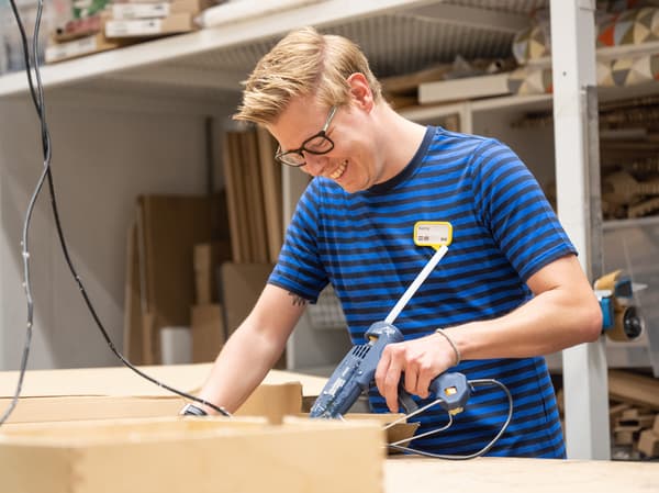 An IKEA co-worker in a blue striped IKEA uniform smile as he works with an electric glue gun in a workshop.