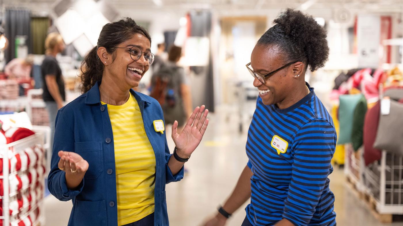 An IKEA co-worker in uniform and carrying a yellow IKEA bag, coming down IKEA store stairs to greet customers.
